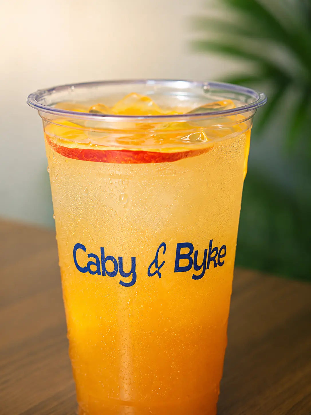 A close-up of a frothy coffee drink in a clear plastic cup with the blue text 'Caby & Byke' on the front, topped with whipped cream and cocoa powder, sitting on a wooden surface.