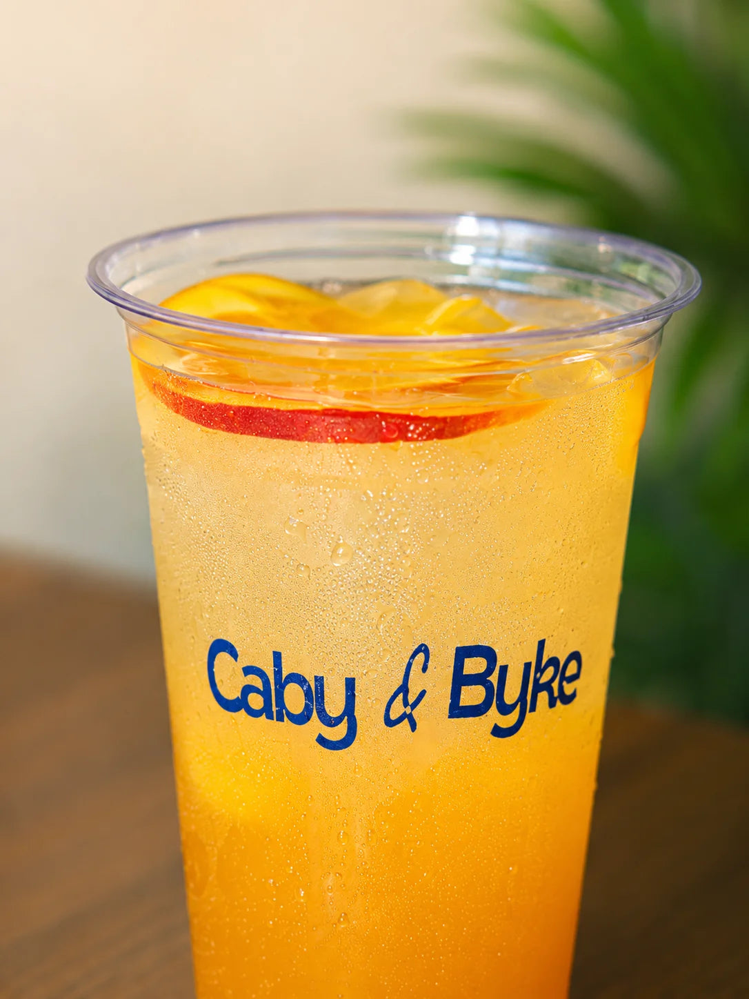 A clear plastic cup with condensation, filled with an orange-yellow beverage and garnished with a red slice and yellow fruit, printed with the blue text 'Caby & Byke'.