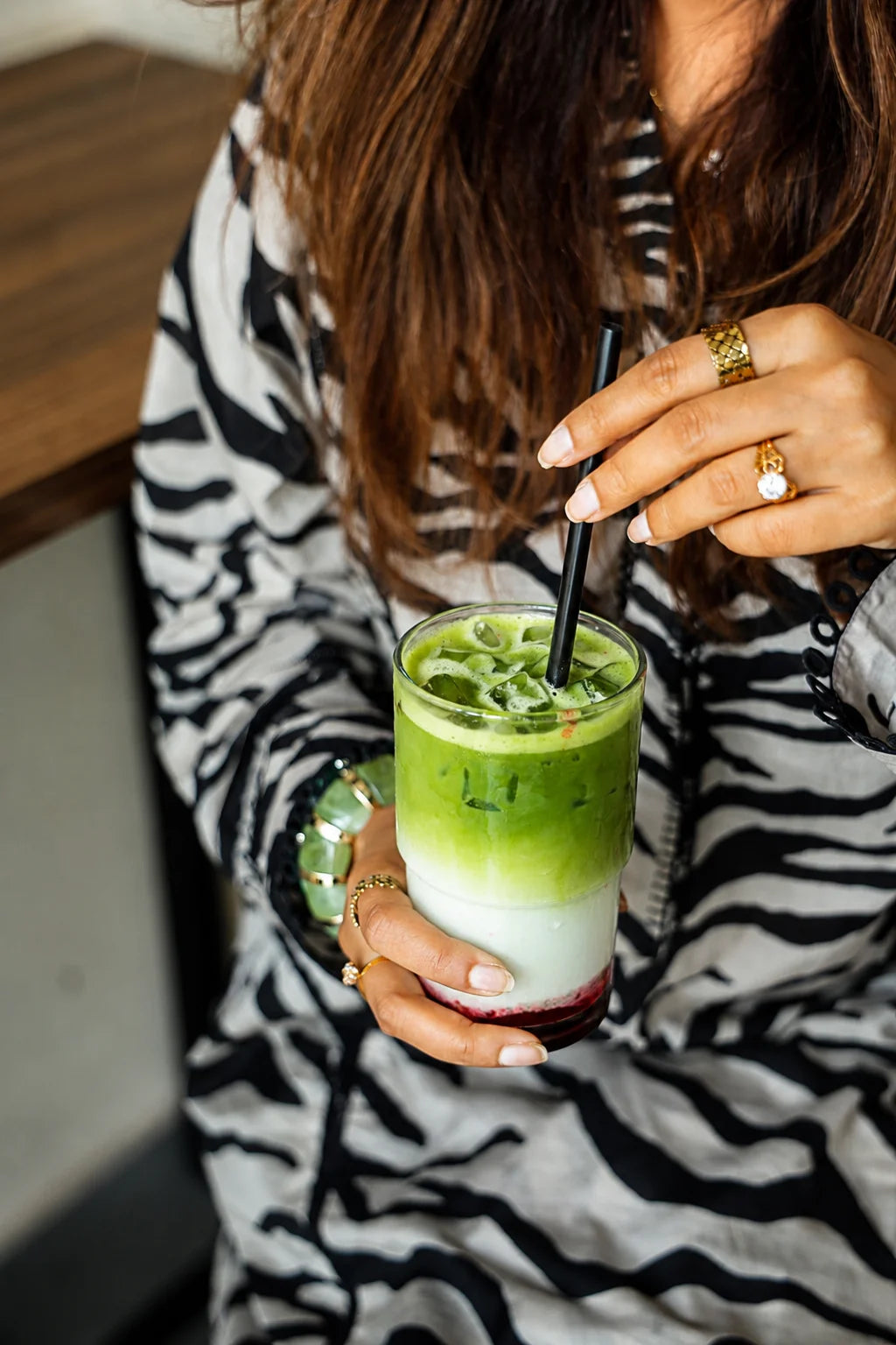 A person in a black and white zebra-print top holds an iced matcha latte with a black straw. The person has long brown hair, gold rings, and a green bracelet, and is holding the drink in a cafe setting.