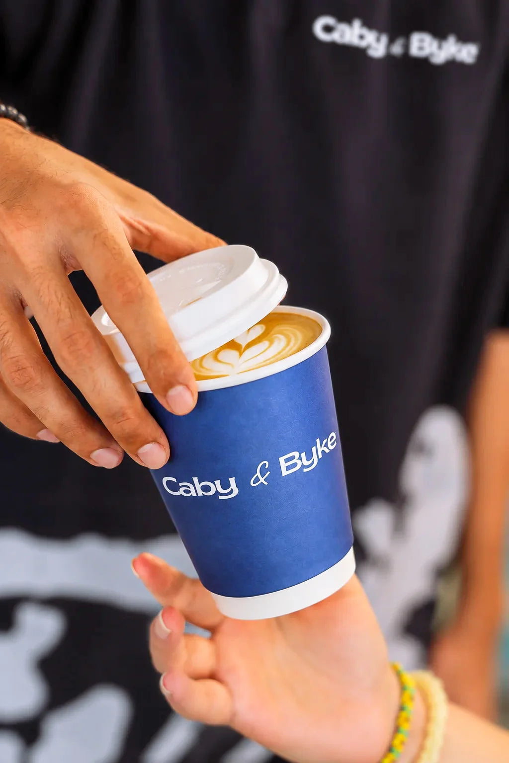 A close-up of a person handing a blue paper coffee cup with a white lid to another person. The cup is branded 'Caby & Byke' in white text, and the background shows a person wearing a matching black branded shirt.