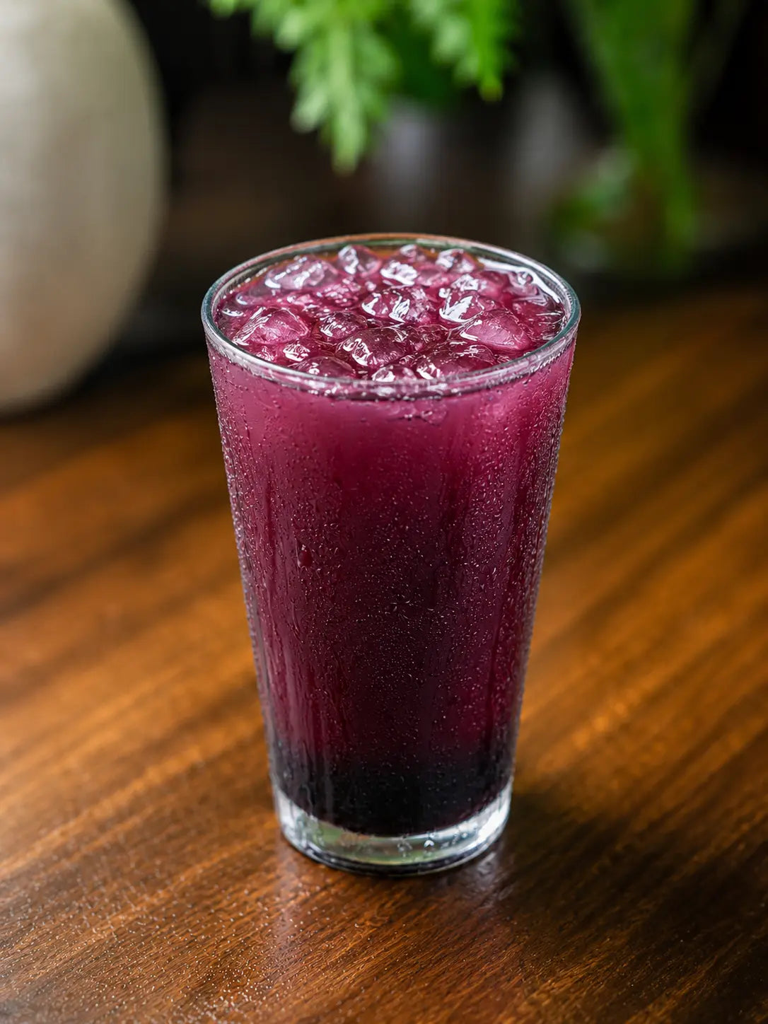 A clear glass tumbler filled with a deep purple berry drink and crushed ice, set on a dark wooden table with a softly blurred background.