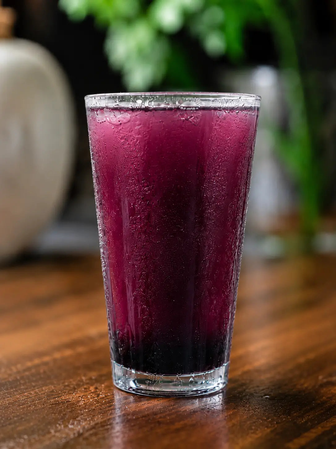 A tall clear glass filled with a deep purple-red beverage, likely iced berry juice, with condensation on the outside, set on a wooden table with a blurred background.